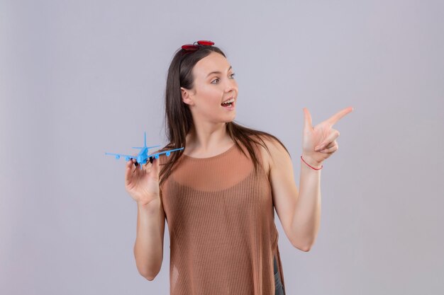 Junge schöne Frau mit roter Sonnenbrille auf Kopf, der Spielzeugflugzeug hält, das mit Finger zur Seite positiv und glücklich steht über weißem Hintergrund zeigt