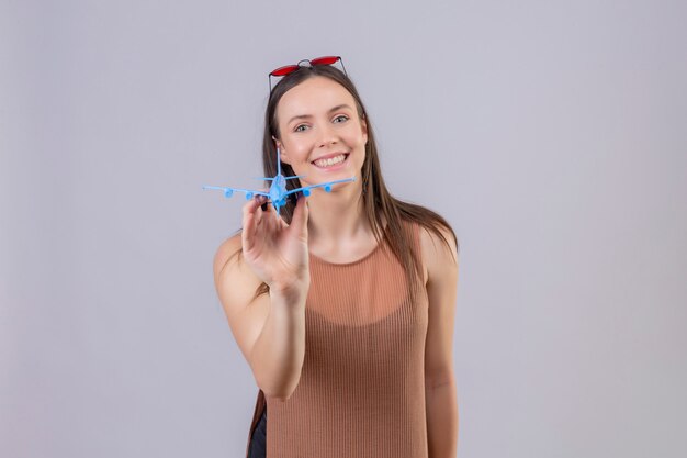 Junge schöne Frau mit roter Sonnenbrille auf Kopf, der Spielzeugflugzeug hält, das mit glücklichem Gesicht über weißer Wand lächelt