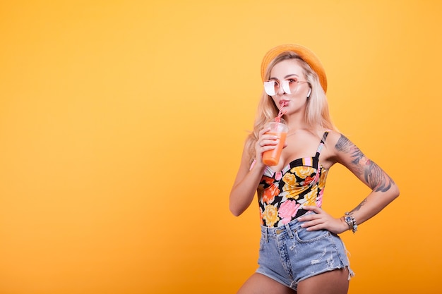 Junge schöne Frau trinkt frischen Orangensaft mit Hut und Sonnenbrille im Studio