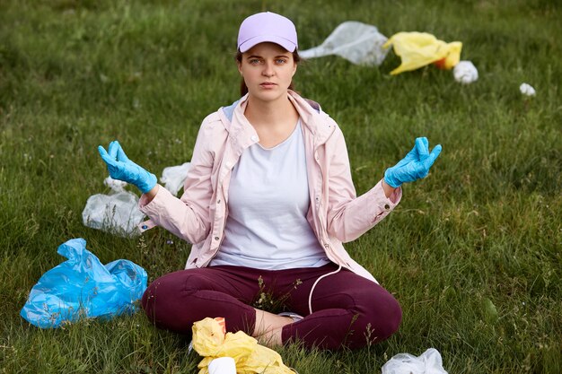 Junge schöne Freiwillige, die Müll im Park aufheben, müde und wütend sein, versuchen, sich zu entspannen, in Lotushaltung auf Gras sitzen, Kamera betrachten, Freizeitkleidung tragen.