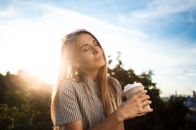 Junge schöne fröhliche Frau, die durch Stadt geht, lächelt und Kaffee hält.