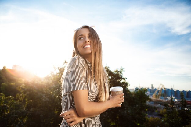 Junge schöne fröhliche Frau, die durch Stadt geht, lächelt und Kaffee hält.