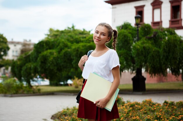 Junge schöne fröhliche Studentin, die lächelt und Ordner draußen hält.