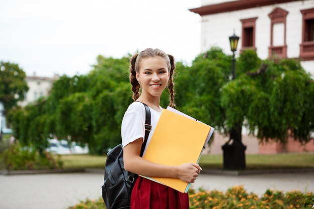 Junge schöne fröhliche Studentin, die lächelt und Ordner draußen hält.