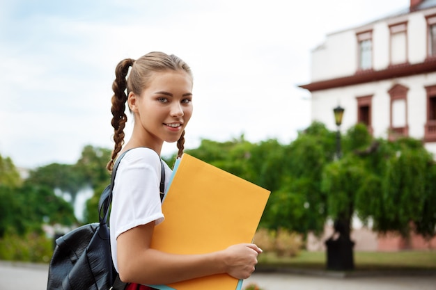 Junge schöne fröhliche Studentin, die lächelt und Ordner draußen hält.