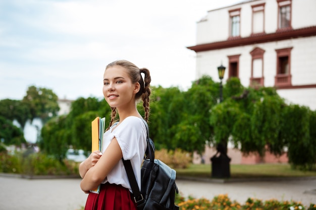 Junge schöne fröhliche Studentin, die lächelt und Ordner draußen hält.