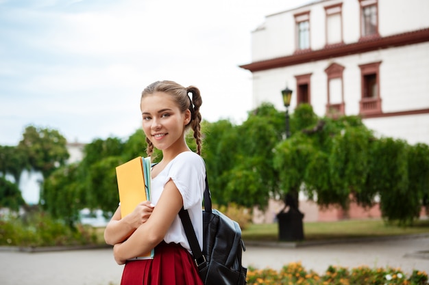 Junge schöne fröhliche Studentin, die lächelt und Ordner draußen hält.