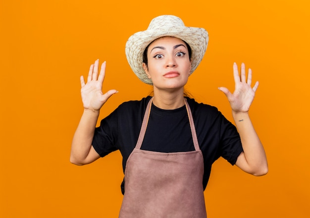 Junge schöne Gärtnerin in der Schürze und im Hut, die Hände in der Übergabe anheben, die überrascht sind, über orange Wand zu stehen