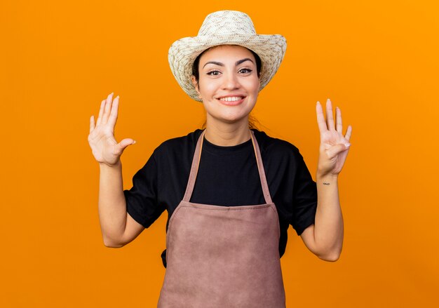 Junge schöne Gärtnerin in Schürze und Hut, die mit den Fingern Nummer neun lächelnd über orange Wand stehend zeigt und zeigt