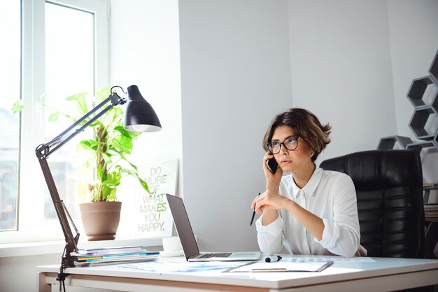 Junge schöne Geschäftsfrau, die am Telefon am Arbeitsplatz im Büro spricht.