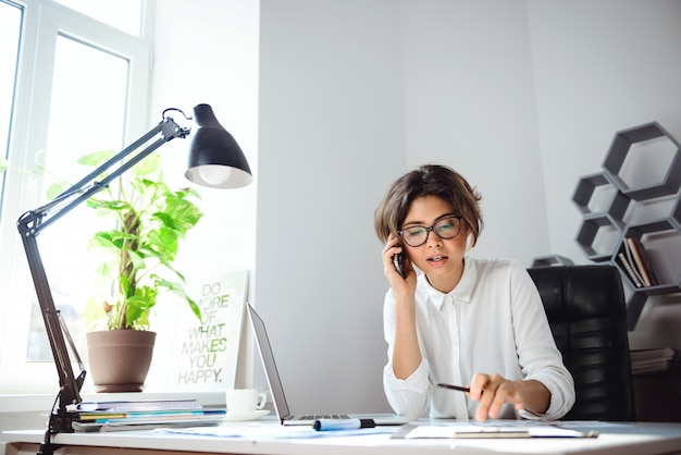 Junge schöne Geschäftsfrau, die am Telefon am Arbeitsplatz im Büro spricht.