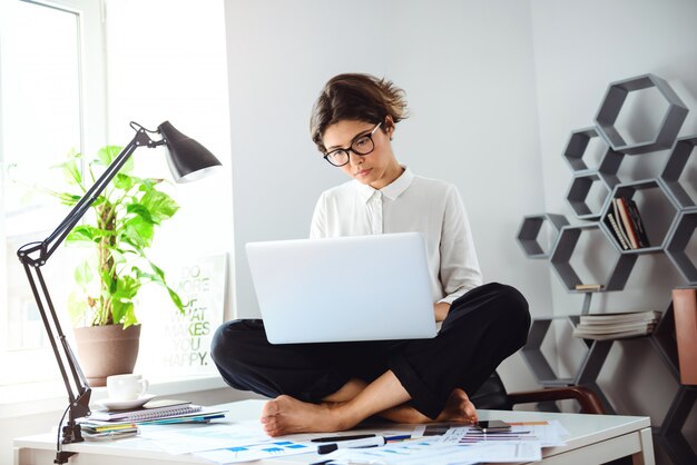 Junge schöne Geschäftsfrau, die auf Tisch mit Laptop am Arbeitsplatz sitzt.