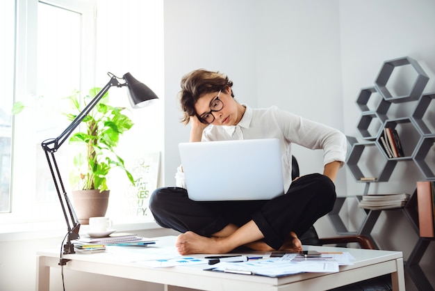 Junge schöne Geschäftsfrau, die auf Tisch mit Laptop am Arbeitsplatz sitzt.