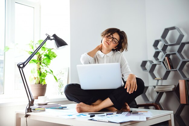 Junge schöne Geschäftsfrau, die auf Tisch mit Laptop am Arbeitsplatz sitzt.