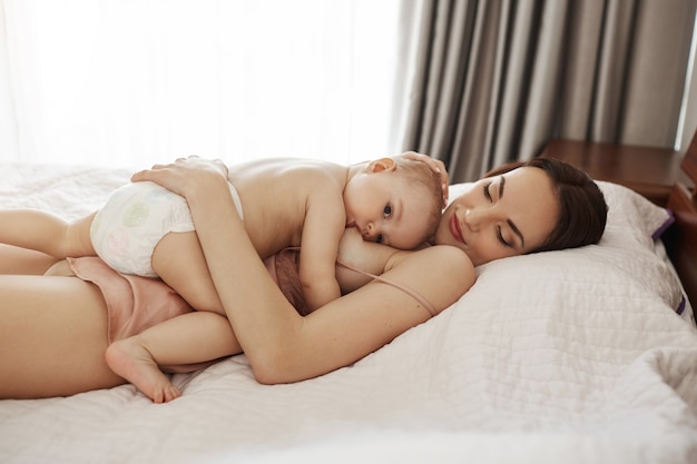 Junge schöne glückliche Mutter, die ihr Baby umarmt, das zu Hause auf dem Bett liegt.