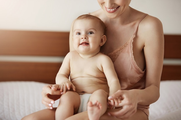 Junge schöne glückliche Mutter in Nachtwäsche und ihr neugeborenes Baby sitzen am Morgen lächelnd auf dem Bett und spielen zusammen.