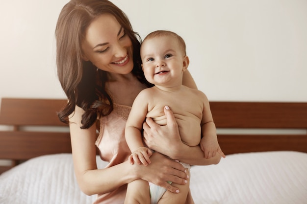 Junge schöne glückliche Mutter in Nachtwäsche und ihr neugeborenes Baby sitzen am Morgen lächelnd auf dem Bett und spielen zusammen