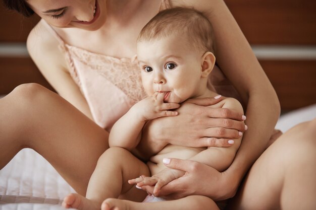 Junge schöne glückliche Mutter in Nachtwäsche und ihr neugeborenes Baby sitzen auf dem Bett lächelnd zusammen zu Hause spielend