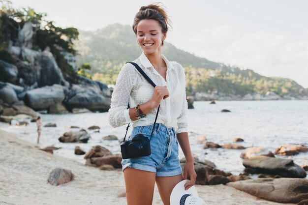Junge schöne Hipster-Frau in den Sommerferien in Asien, entspannend am tropischen Strand, digitale Fotokamera, lässiger Boho-Stil, Seelandschaft, schlank gebräunt, Reisen, lächelnd, glücklich, positiv