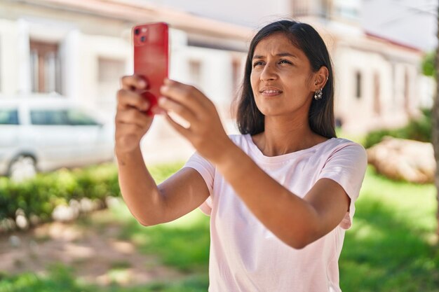 Junge schöne hispanische Frau lächelt selbstbewusst und macht im Park ein Foto per Smartphone