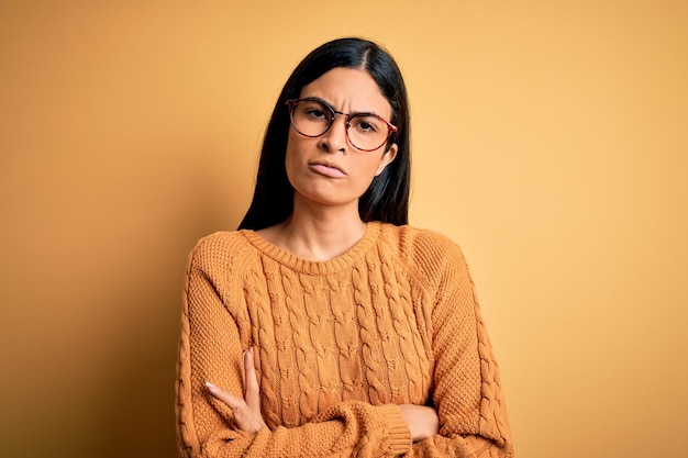 Junge schöne hispanische Frau mit Brille über gelb isoliertem Hintergrund skeptisch und nervös missbilligender Gesichtsausdruck mit verschränkten Armen Negative Person