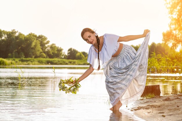 Junge schöne kaukasische Frau, die am Ufer des Flusses steht. Traditionelles Landschaftsbild mit Mädchen im Vordergrund und Kopienraum. Brunette, der einen Kranz hält. Sonneneruption