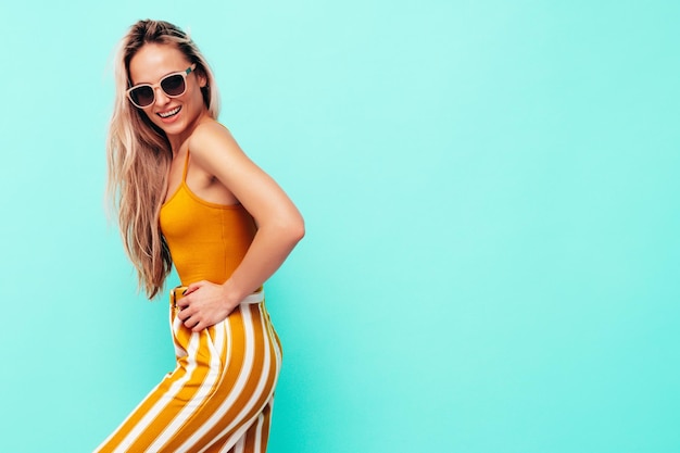 Junge schöne lächelnde blonde Frau in trendiger gelber Sommerkleidung Sexy sorglose Frau posiert in der Nähe der blauen Wand im Studio Positives Modell, das Spaß im Innenbereich hat Fröhlich und glücklich In Sonnenbrille