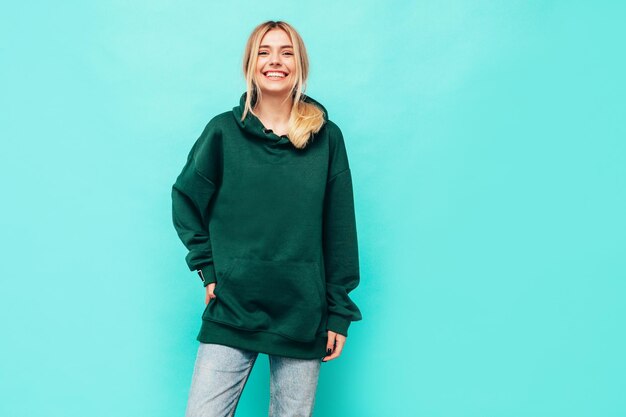 Junge schöne lächelnde Frau im trendigen grünen Hoodie im Sommer Sexy sorglose Frau posiert in der Nähe der blauen Wand im Studio Positives blondes Modell, das Spaß hat und verrückt wird Fröhlich und glücklich