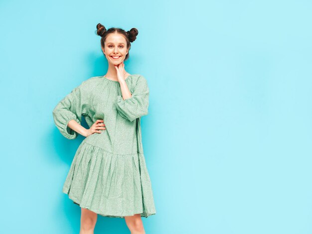Junge schöne lächelnde Frau im trendigen grünen Sommerkleid Sexy sorglose Frau mit Frisur mit zwei Hörnern, die in der Nähe der blauen Wand im Studio posiert Positives Modell, das Spaß hat Fröhlich und glücklich