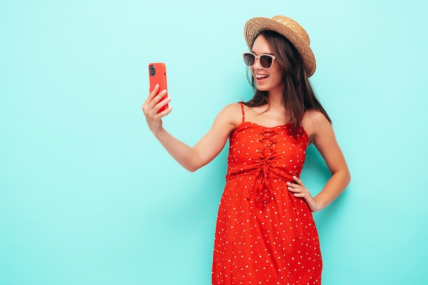 Junge schöne lächelnde Frau im trendigen roten Sommerkleid Sexy sorglose Frau posiert in der Nähe der blauen Wand im Studio Positives brünettes Modell, das Spaß mit Hut und Sonnenbrille hat Selfie-Fotos machen