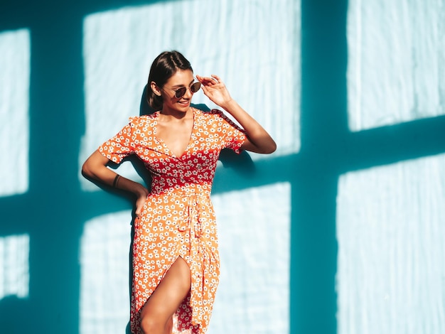 Junge schöne lächelnde Frau im trendigen roten Sommerkleid Sexy sorglose Frau posiert in der Nähe der blauen Wand im Studio Positives Modell, das Spaß hat Fröhlich und glücklich Am sonnigen Tag Schatten vom Fenster