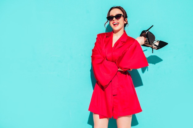 Kostenloses Foto junge schöne lächelnde frau im trendigen roten sommerkleid sexy sorglose frau posiert in der nähe der blauen wand im studio positives modell, das spaß hat fröhlich und glücklich hält ihre hochhackigen schuhe in den händen