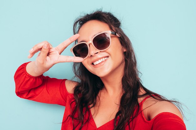 Junge schöne lächelnde Frau im trendigen roten Sommertop und Jeanskleidung Sexy sorglose Frau posiert in der Nähe der blauen Wand im Studio Positives brünettes Modell, das Spaß hat Pov-Selfie mit Sonnenbrille aufnehmen