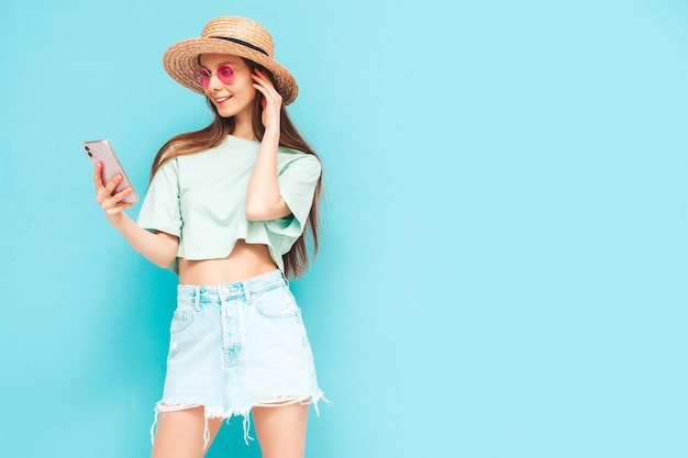 Junge schöne lächelnde Frau im trendigen Sommerrock Sexy Frau mit Posing in der Nähe der gelben Wand im Studio Positives Modell, das Spaß hat In Sonnenbrille und Hut Blick auf Smartphone mit Apps