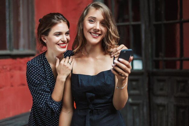 Junge, schöne, lächelnde Frau in schwarzem Kleid, die ein Handy in der Hand hält und glücklich mit einem fröhlichen Freund in der Nähe des alten Hofes in die Kamera schaut