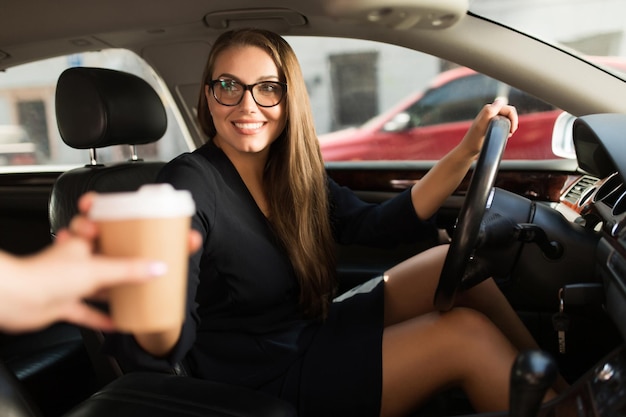 Junge schöne lächelnde Frau in schwarzem Kleid und Brille, die hinter dem Lenkrad im Auto sitzt, während sie glücklich Kaffee zum Mitnehmen nimmt