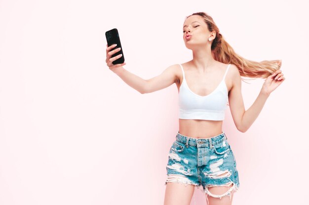 Junge schöne lächelnde Frau in trendigen Sommer-Jeans-Shorts Sexy sorglose blonde Frau posiert in der Nähe der Wand im Studio Positives Modell, das Spaß hat Fröhlich und glücklich in Sonnenbrillen IsolatedTaking selfie