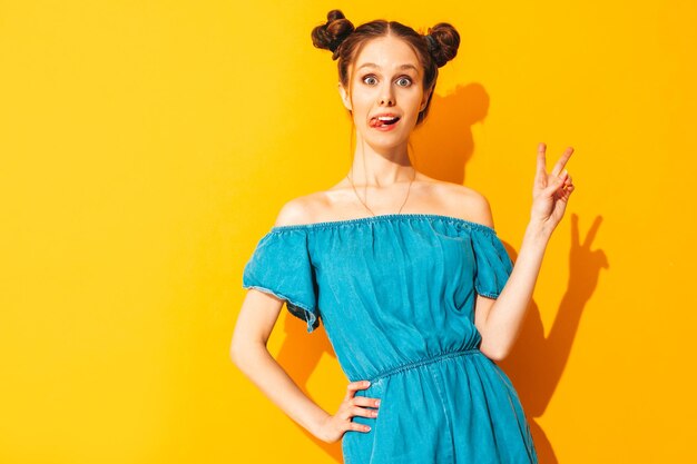 Junge schöne lächelnde Frau in trendigen Sommerjeans-Overalls Sexy sorglose Frau mit zwei Hörnern Frisur posiert in der Nähe der gelben Wand im Studio Positives Modell, das Spaß hat Isoliert