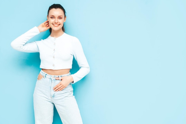 Junge schöne lächelnde Frau in trendigen Sommerjeans und Top-Kleidung Sexy sorglose Frau posiert in der Nähe der blauen Wand im Studio Positives brünettes Modell, das Spaß hat Fröhlich und glücklich