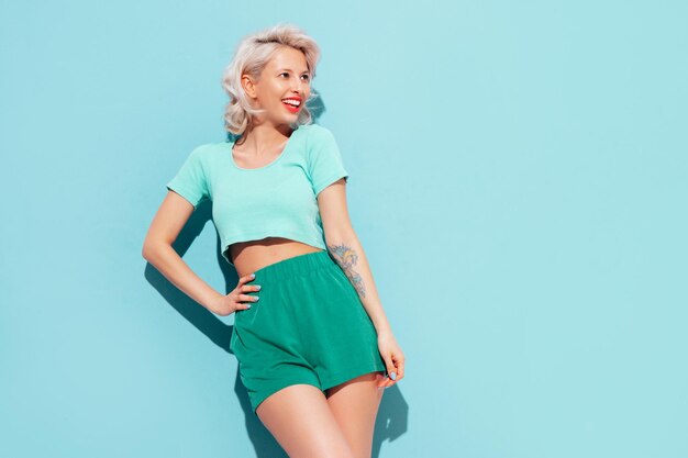 Junge schöne lächelnde Frau in trendigen Sommershorts und Top-Kleidung Sexy sorglose Frau posiert in der Nähe der blauen Wand im Studio Positives blondes Modell, das Spaß hat und verrückt wird Fröhlich und glücklich