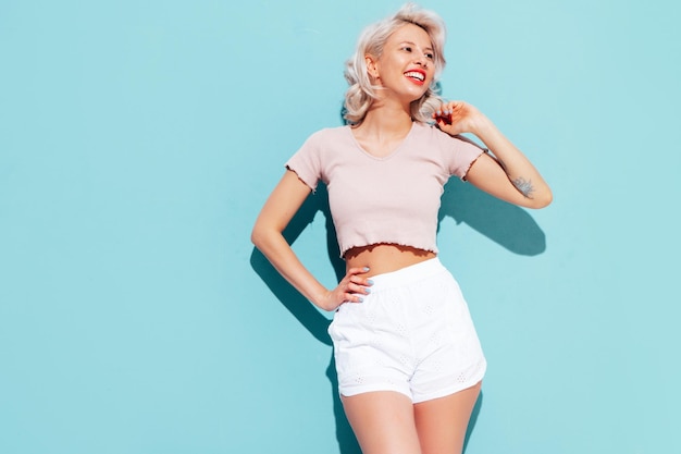 Junge schöne lächelnde Frau in trendigen Sommershorts und Top-Kleidung Sexy sorglose Frau posiert in der Nähe der blauen Wand im Studio Positives blondes Modell, das Spaß hat und verrückt wird Fröhlich und glücklich