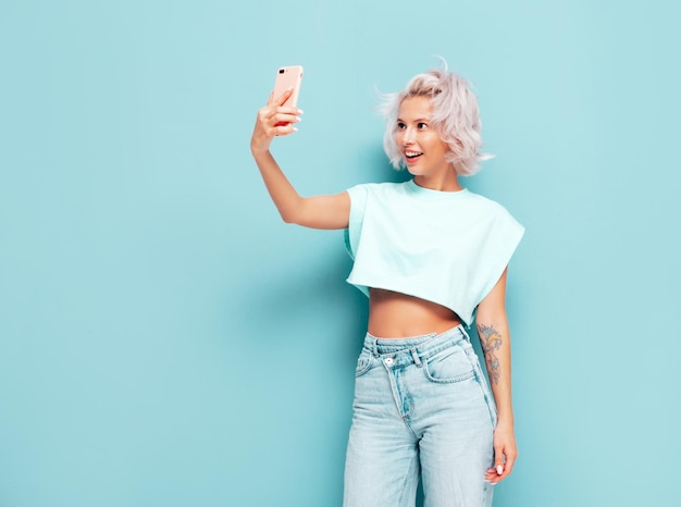 Junge schöne lächelnde Frau in trendiger Sommerkleidung Sexy sorglose Frau posiert in der Nähe der blauen Wand im Studio Positives blondes Modell, das Spaß hat und verrückt wird Fröhlich und glücklich Selfie-Fotos machen