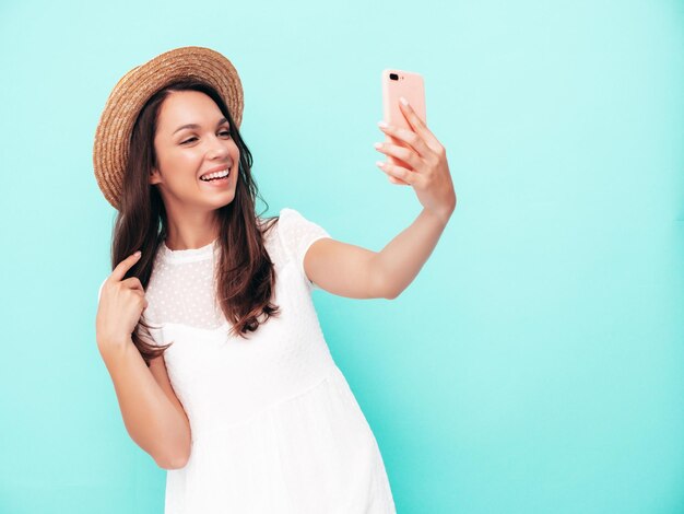 Junge schöne lächelnde Frau in trendiger Sommerkleidung Sexy sorglose Frau posiert in der Nähe der blauen Wand im Studio Positives brünettes Modell, das Spaß hat Fröhlich und glücklich Nimmt Selfie mit Hut