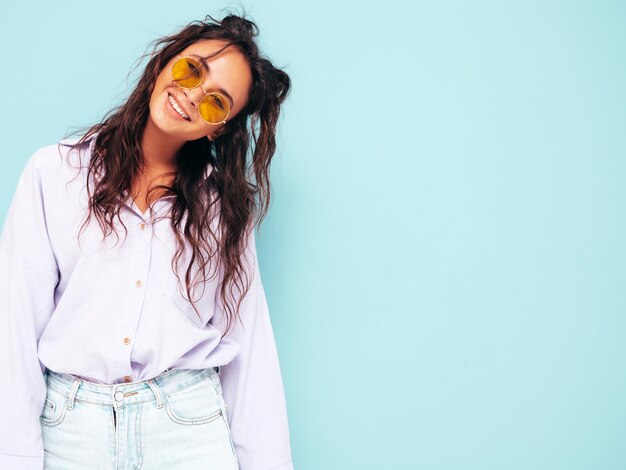 Junge schöne lächelnde Frau in trendiger Sommerkleidung Sexy sorglose Frau posiert in der Nähe der blauen Wand im Studio Positives brünettes Modell, das Spaß hat und verrückt wird Fröhlich und glücklich mit Sonnenbrille