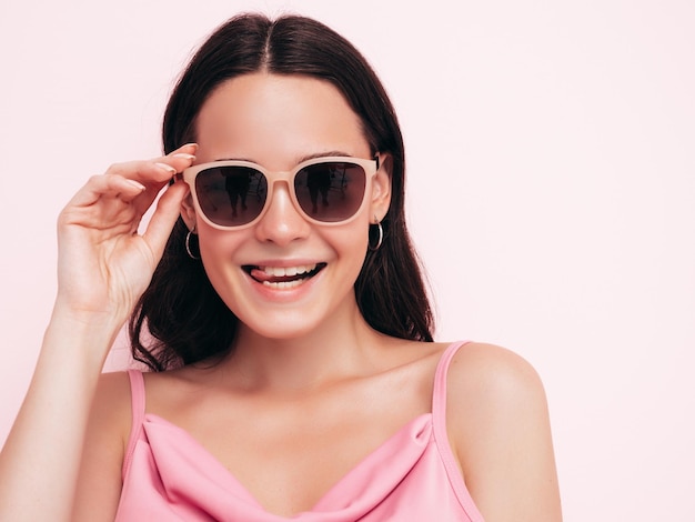 Junge schöne lächelnde Frau in trendiger Sommerkleidung Sexy sorglose Frau posiert in der Nähe von rosa Wand im Studio Positives Modell, das Spaß im Innenbereich hat Sonnenbrille abziehen Zeigt Zunge