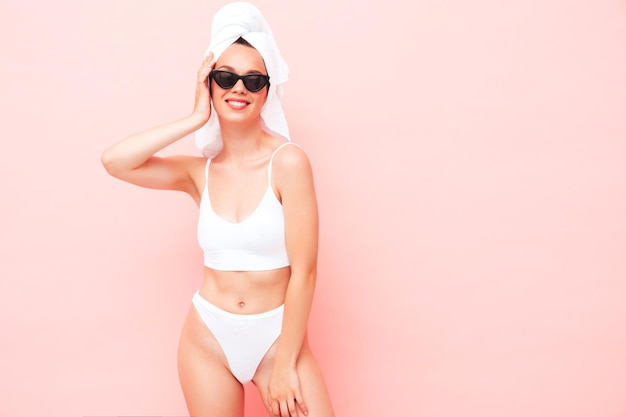 Junge schöne lächelnde Frau in weißen Dessous Sexy sorgloses Modell in Unterwäsche und Handtuch auf dem Kopf posiert rosa Wand im Studio Positive und glückliche Frau, die den Morgen mit Sonnenbrille genießt