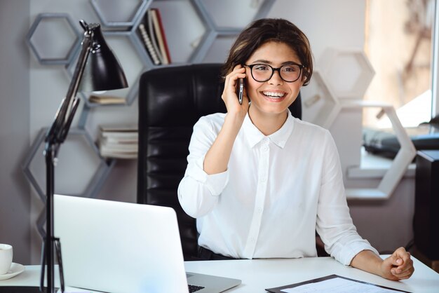 Junge schöne lächelnde Geschäftsfrau, die am Telefon am Arbeitsplatz im Büro spricht.