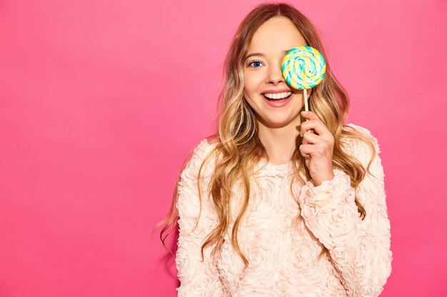 Junge schöne lächelnde Hippie-Frau in der modischen Sommerkleidung. Sexy sorglose Frau, die nahe rosa Wand aufwirft. Positives Modell, das ihr Auge durch Lutscher versteckt