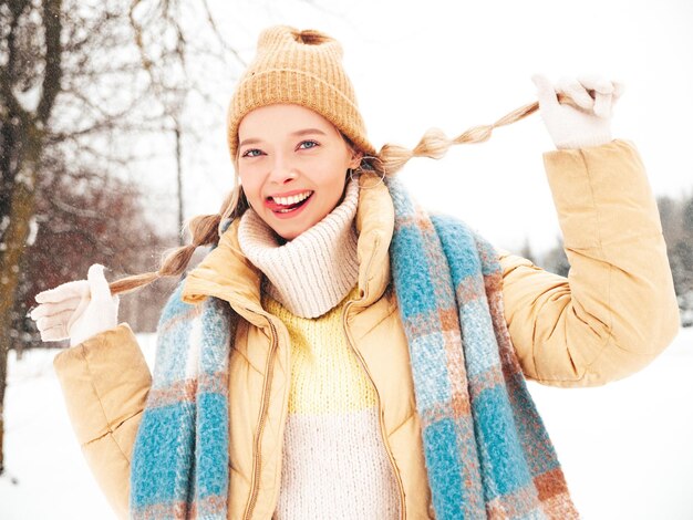 Junge schöne lächelnde Hippie-Frau in trendiger warmer Kleidung und Schal. Sorglose Frau, die auf der Straße im Park posiert. Positives reines Modell, das Spaß im Schnee hat. Wintermomente genießen. Zeigt Zunge