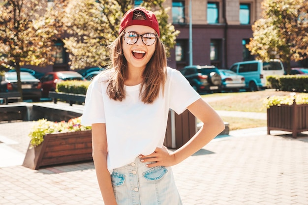 Junge schöne lächelnde Hipster-Frau im trendigen weißen Sommert-shirt Sexy sorglose Frau, die bei Sonnenuntergang auf dem Straßenhintergrund mit Mütze posiert Positives Modell im Freien Fröhlich und glücklich